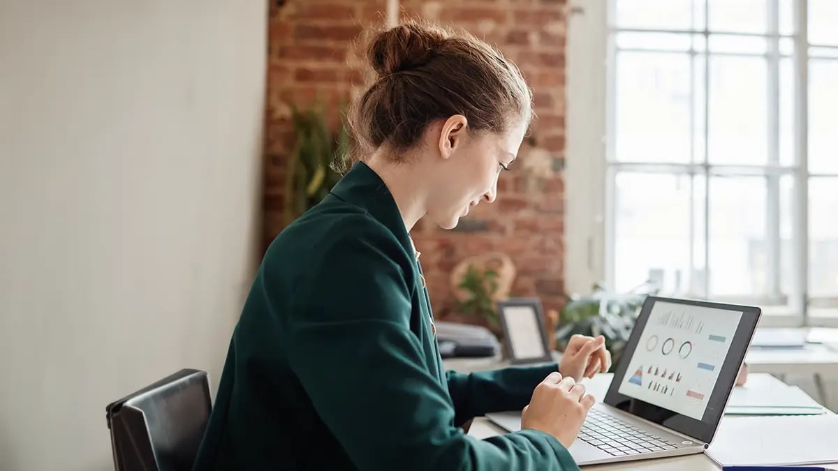 A woman in a green blazer sat at a desk looking at a laptop screen showing graphs and charts.