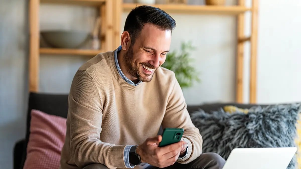 Man holding a phone in his right hand, looking at a laptop and smiling.