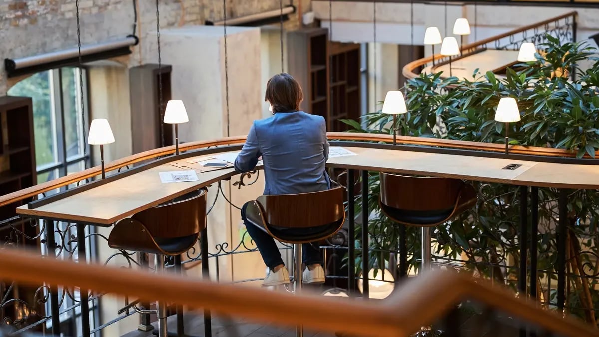 A man sitting on the middle out of three chairs at a corner long desk overlooking the lower floor in a hotel lobby.