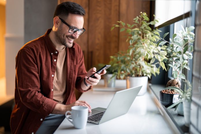 Satisfied modern millennial man in stylish casual clothes using smartphone and laptop computer