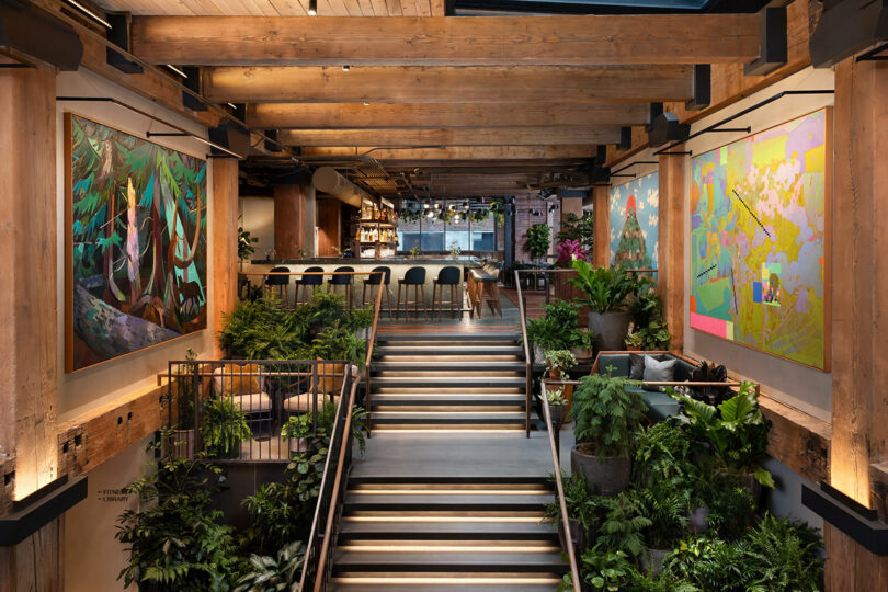 Modern interior with wooden beams, large colorful paintings, lush green plants, and a central staircase leading to a bar area with high stools in the background.