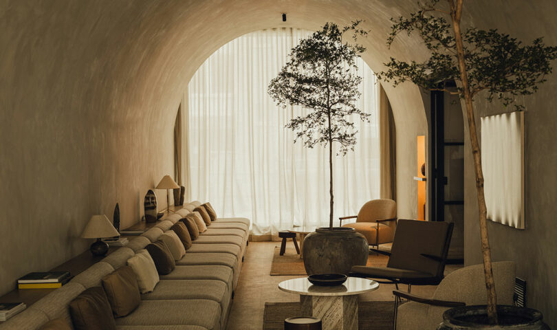 A modern living room with a vaulted ceiling, long beige sofa, armchairs, potted trees, round coffee table, and floor-to-ceiling sheer curtains letting in natural light.