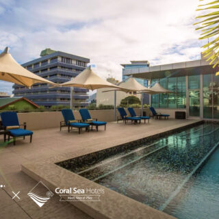 Rooftop pool area with blue lounge chairs and beige umbrellas, glass-walled building, palm fronds, and a sunny sky; hotel branding logos appear in the bottom corner.