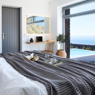 Bright seaside bedroom with white linens, a gray patterned throw, and a desk facing a large window overlooking the ocean and a pool.