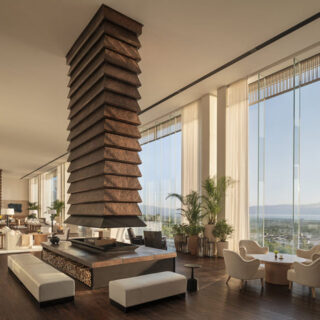 Spacious modern hotel lounge with a tall stacked stone fireplace, seating areas, and floor-to-ceiling windows overlooking the landscape.