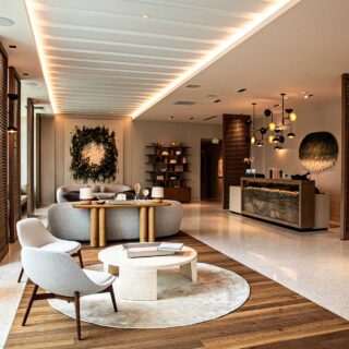Bright hotel lobby with curved gray sofas around a round wooden table, large windows, and warm ambient lighting.