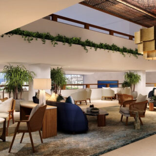 Spacious hotel lounge with cream sofas, wooden chairs, round tables, green plants, and a geometric pendant light.