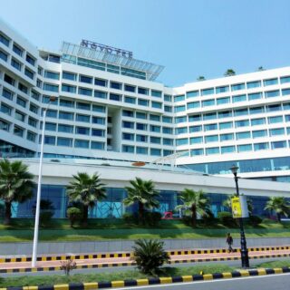Exterior view of a modern white hotel with a grid of blue-tinted windows and a NOVOTEL sign on the roof, palm trees in front.