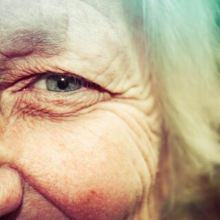 Close-up of an elderly person's eye and surrounding wrinkled skin, showing blue-gray iris and fine facial lines.