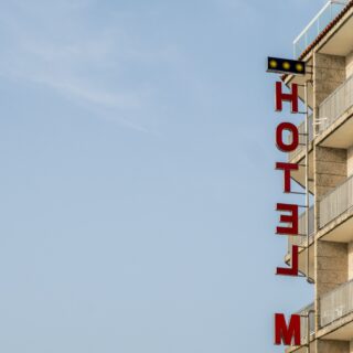 Red vertical letters spell 'HOTEL' on the side of a beige apartment building under a blue sky.