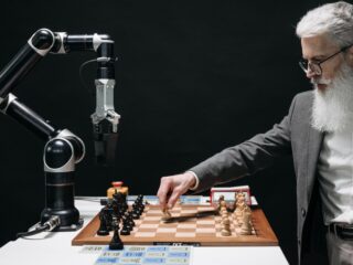 Bearded older man in a suit playing chess opposite a robotic arm in a dark studio setting.