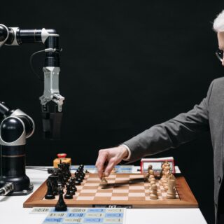 Bearded older man in a suit playing chess opposite a robotic arm in a dark studio setting.