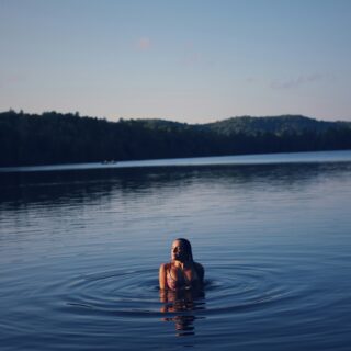 Person swimming in a large lake with a forested shoreline and clear blue sky in the background.
