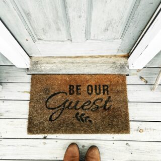 Top-down view of a brown welcome mat on weathered white wooden steps, reading 'Be Our Guest' in script, with brown shoes at the bottom edge.