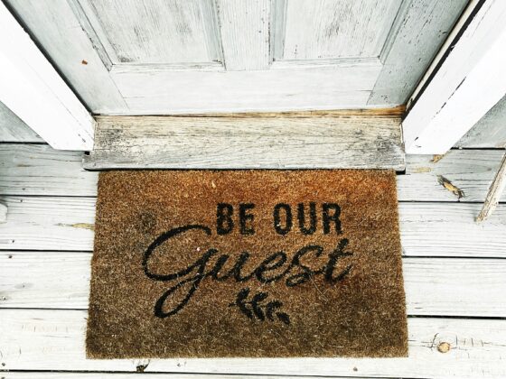 Top-down view of a brown welcome mat on weathered white wooden steps, reading 'Be Our Guest' in script, with brown shoes at the bottom edge.