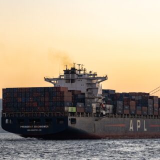 Container ship loaded with stacked shipping containers gliding on the water with a city skyline and bridge in the distance at sunset.