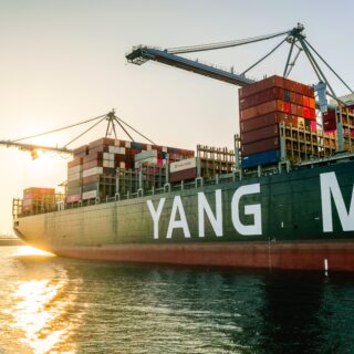Large cargo ship loaded with colorful containers at a port, cranes overhead during sunset.