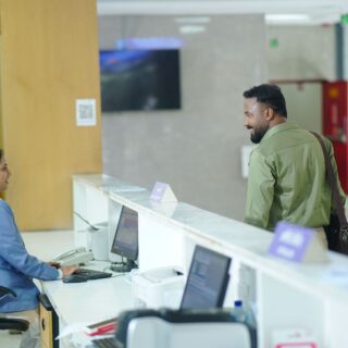 Teller in a blue blazer sits at a computer desk as a male customer stands at the counter, talking in a bright lobby.