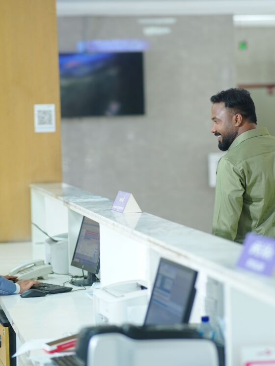 Teller in a blue blazer sits at a computer desk as a male customer stands at the counter, talking in a bright lobby.