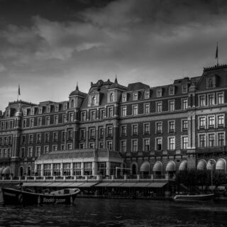 Black-and-white view of a grand riverside hotel along a canal, with boats moored in the foreground and a cloudy sky above.