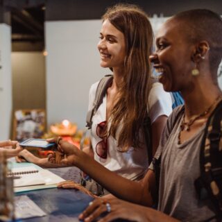 Two women smiling at a counter as one hands a card to a staff member in a shop or reception area.
