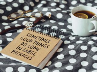 Notebook on a polka-dot blanket with the phrase “Sometimes good things come in large packages,” beside a cup of tea with a lemon slice and glasses nearby.
