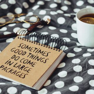 Notebook on a polka-dot blanket with the phrase “Sometimes good things come in large packages,” beside a cup of tea with a lemon slice and glasses nearby.