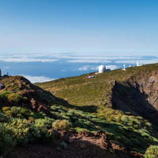 Mountain ridge with white observatories and domes overlooking a sea of clouds under a clear blue sky, late afternoon light.