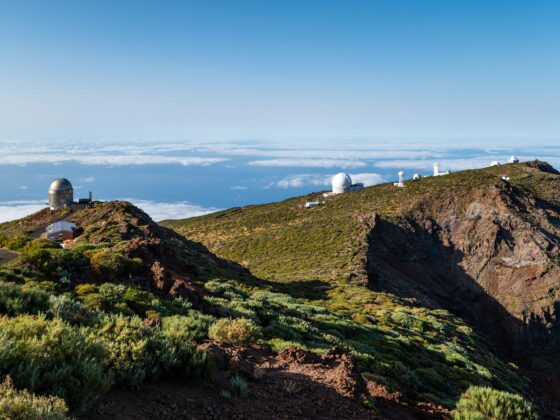Mountain ridge with white observatories and domes overlooking a sea of clouds under a clear blue sky, late afternoon light.