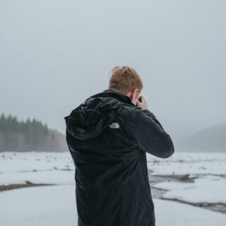 Person in a black jacket photographed from behind, holding a camera in a snowy, overcast landscape with distant trees.