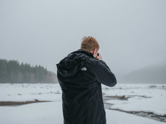 Person in a black jacket photographed from behind, holding a camera in a snowy, overcast landscape with distant trees.