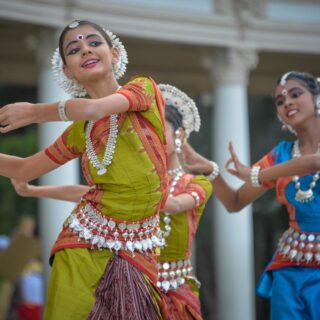 Two female dancers in bright traditional Indian costumes with silver jewelry perform outdoors near columned architecture.