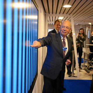 Man in a suit gestures toward a blue wall exhibit as photographers and attendees watch in the background.