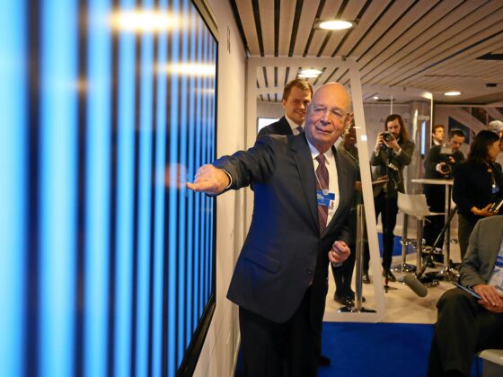 Man in a suit gestures toward a blue wall exhibit as photographers and attendees watch in the background.