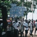 Crowded street with a wooden sign listing hotel, conference hall, wedding hall, swimming pool and playground; a vendor carries a basket of drinks under a tree.