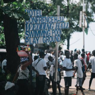 Crowded street with a wooden sign listing hotel, conference hall, wedding hall, swimming pool and playground; a vendor carries a basket of drinks under a tree.