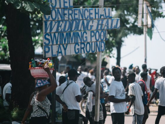 Crowded street with a wooden sign listing hotel, conference hall, wedding hall, swimming pool and playground; a vendor carries a basket of drinks under a tree.