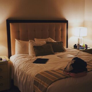 Hotel room at night featuring a queen-size bed with a tufted headboard, two lit nightstand lamps, and cluttered surfaces on the nightstands and bed.