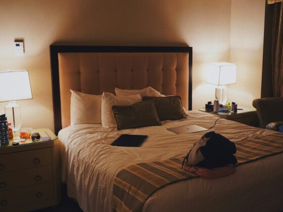 Hotel room at night featuring a queen-size bed with a tufted headboard, two lit nightstand lamps, and cluttered surfaces on the nightstands and bed.