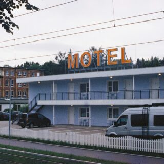 Motel with a large illuminated MOTEL sign on a blue building, a white water tower to the left, and a row of parked cars behind a white picket fence.