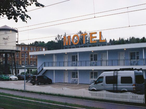 Motel with a large illuminated MOTEL sign on a blue building, a white water tower to the left, and a row of parked cars behind a white picket fence.