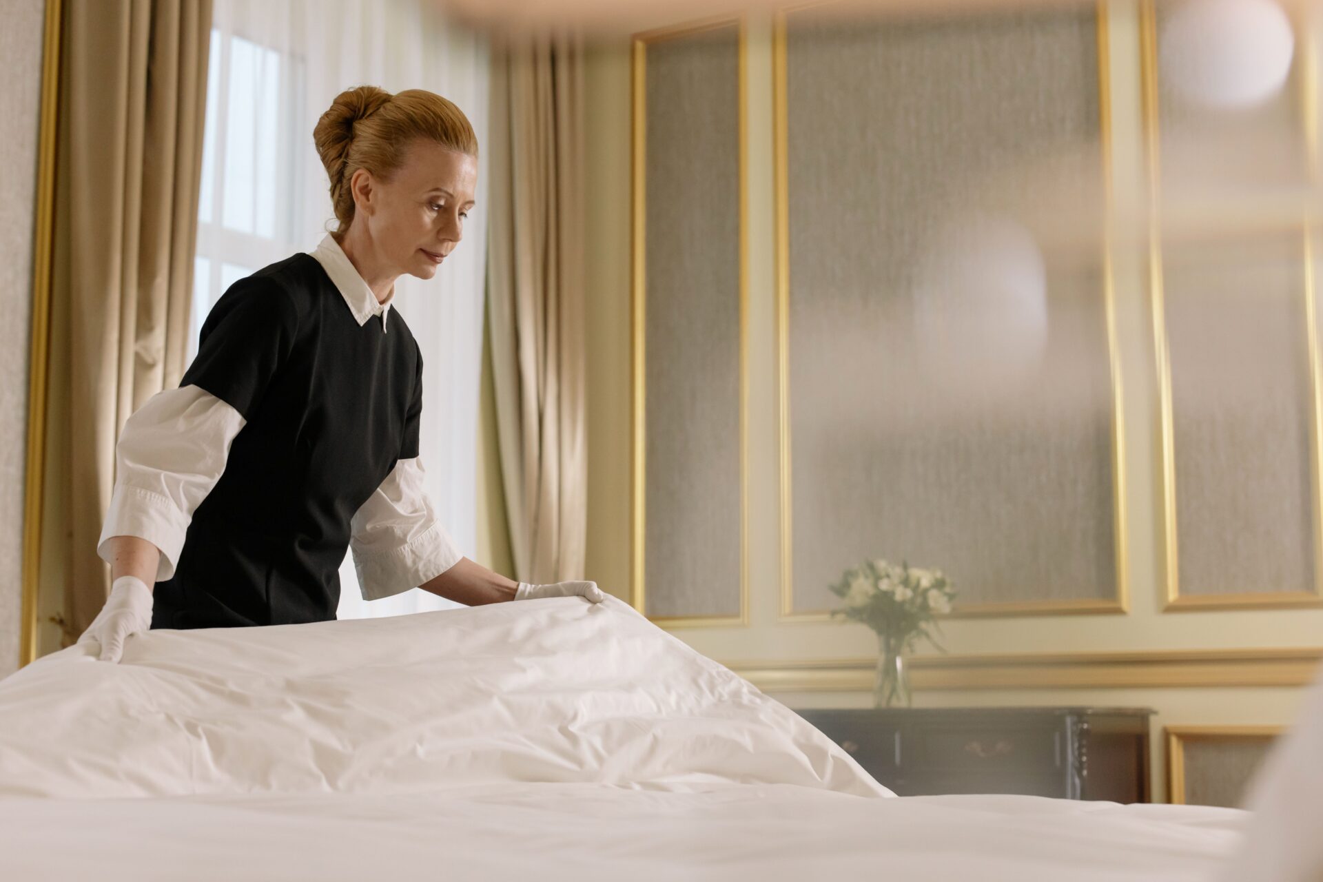 A staff member wearing gloves and a black uniform irons a white sheet on a bed/table in a bright, formal room with gold-framed mirrors in the background.