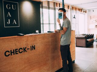 Man at a check-in counter holds a small framed card in front of his face, with 'CHECK-IN' on the wooden desk and a lounge area in the background.