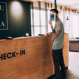 Man at a check-in counter holds a small framed card in front of his face, with 'CHECK-IN' on the wooden desk and a lounge area in the background.