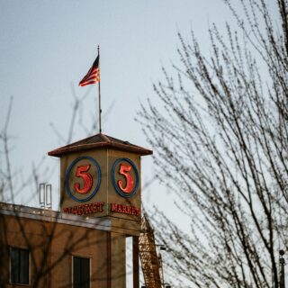 Beige market building with neon blue-outlined '5' signs and the word 'MARKET' on the facade; a flagpole with an American flag atop the roof, framed by bare tree branches against a pale blue sky.
