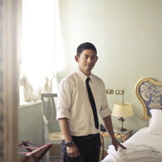 Man in a white dress shirt and black tie stands beside a bed in a hotel room with a blue and gold headboard.