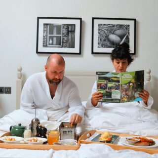Two people in white robes sit up in bed, eating breakfast from a large wooden tray with cups and plates in front of them.