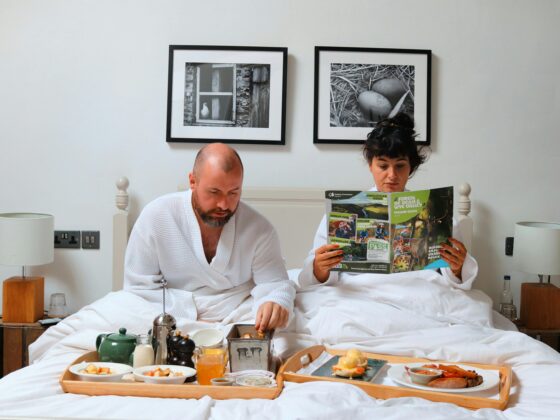 Two people in white robes sit up in bed, eating breakfast from a large wooden tray with cups and plates in front of them.