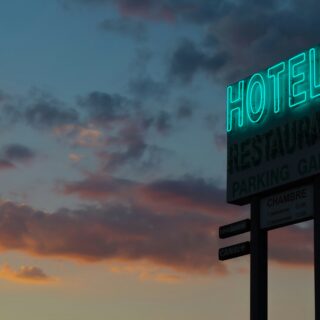 Neon turquoise 'HOTEL' sign on a tall roadside sign against a dusky sunset sky; restaurant and parking panels visible below.