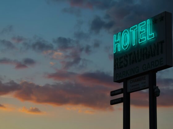 Neon turquoise 'HOTEL' sign on a tall roadside sign against a dusky sunset sky; restaurant and parking panels visible below.